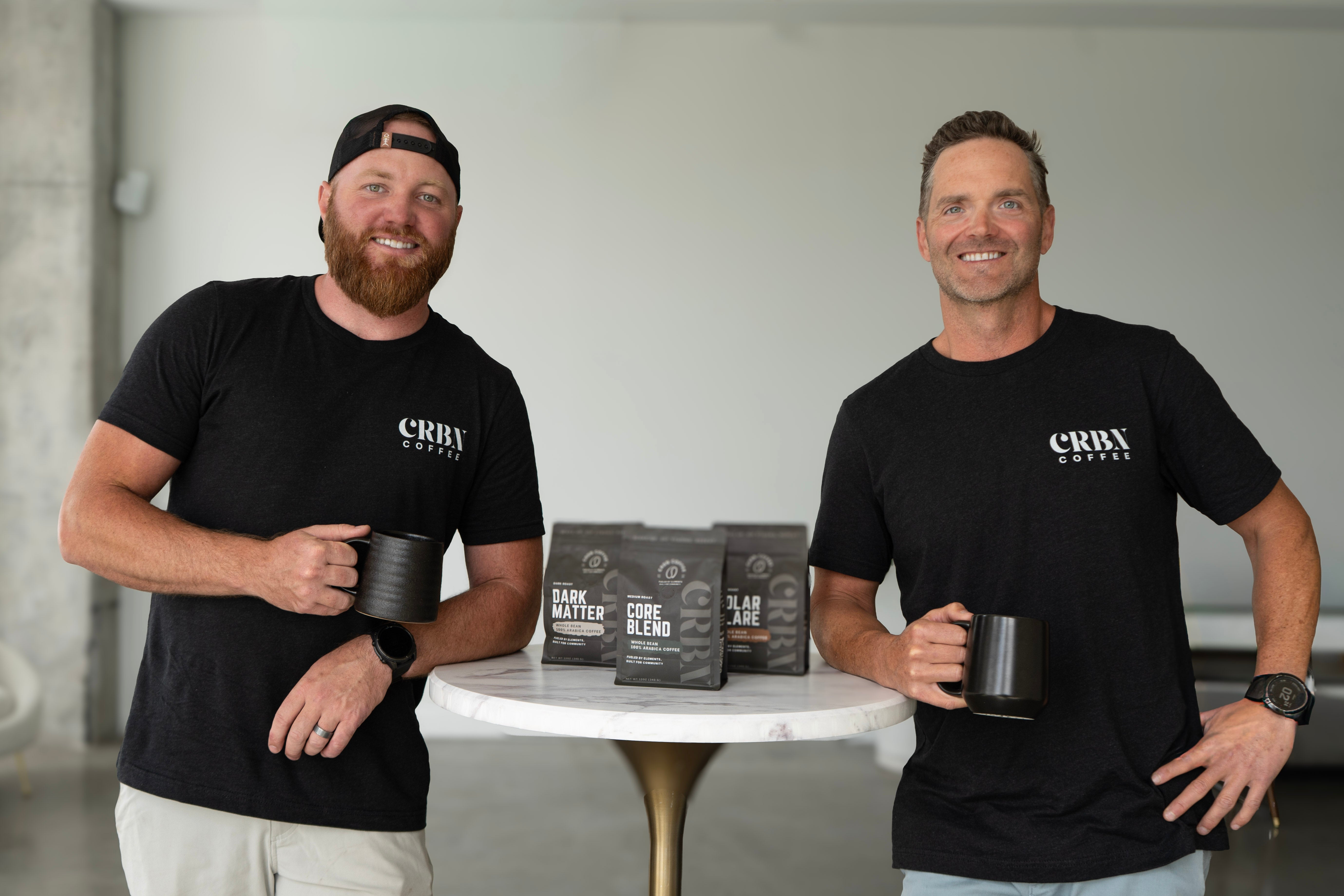 Two men in black t-shirts with 'CRBBY' branding holding coffee cups, standing behind a table with coffee bags.